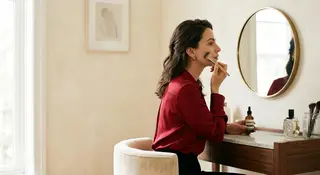 A woman in a cherry-red blouse applying foundation with a brush at a round vanity mirror